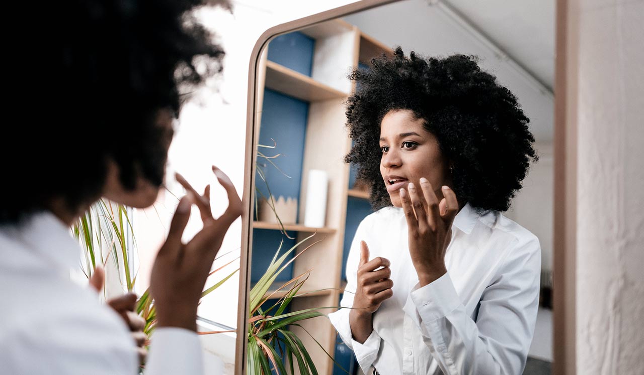 A woman looks at her reflection in the mirror as she applies skin care products.