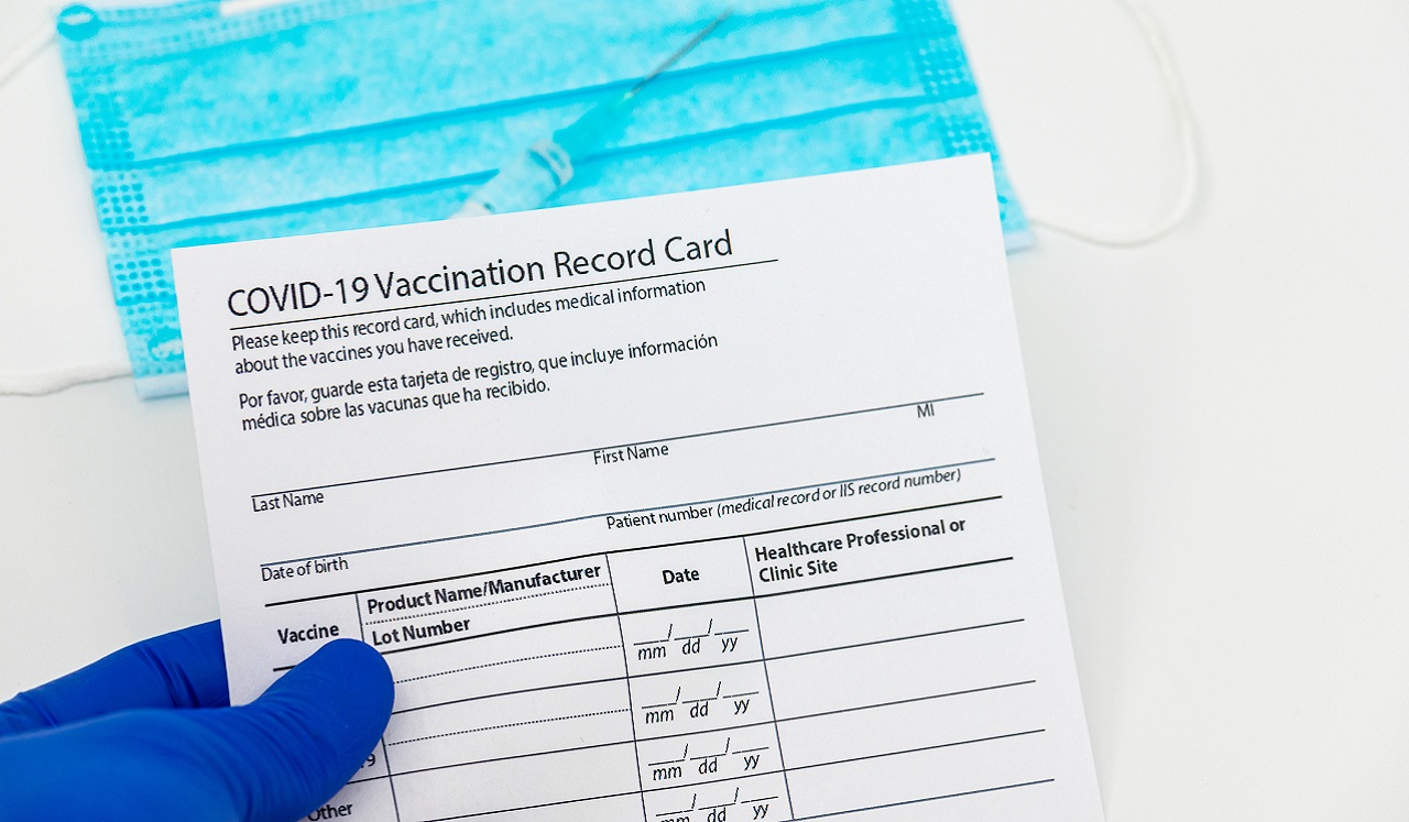 A gloved hand holds a vaccination record card; in the background is a syringe and a medical face mask.